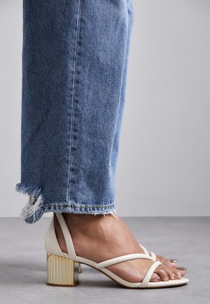 Sandale blanche à talons hauts avec des lanières et un talon carré texturé doré, portée avec un jean en denim bleu effiloché. Gros plan sur le pied et la chaussure.
