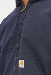 Mørkeblå lynlås hoodie med en frontlomme, der har snore og en guld lynlås. Indeholder et synligt orange logo mærke på lommen.