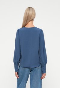 Blå langermet bluse med rund hals, strukturert stoff og knapper på mansjettene. Bak har den rynker på skuldrene og en avslappet passform.