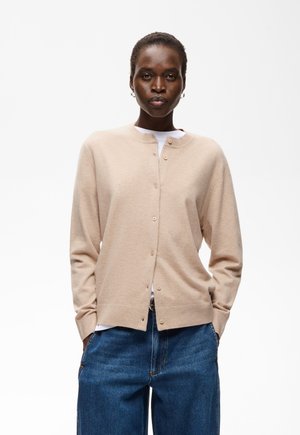Femme portant un cardigan beige boutonné sur une chemise blanche, les mains dans les poches d'un jean bleu, debout devant un fond blanc.