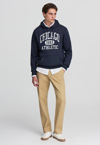 Felpa navy con stampa "CHICAGO 1983 ATHLETIC", tasca a marsupio, abbinata a pantaloni beige e sneaker bianche, creando un outfit casual.