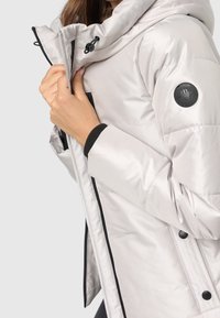 Ljusgrå pufferjacka med huva, med svarta accenter, en logotypapplikation på ärmen och exponerade dragkedjor fram.