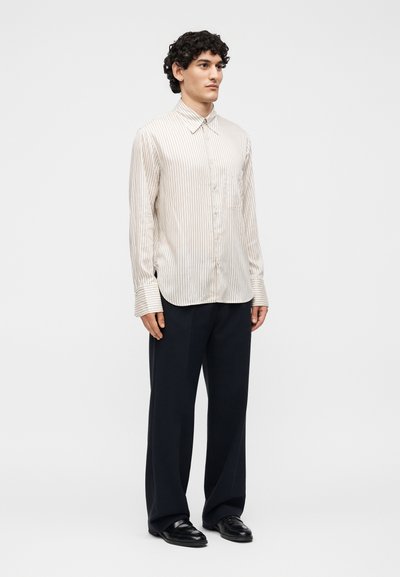 Άνδρας με κατσαρά μαλλιά που φοράει ένα κρεμ πουκάμισο με ρίγες και κουμπιά, μαύρο παντελόνι με φαρδιά πόδια και μαύρα δερμάτινα παπούτσια, στέκεται μπροστά σε λευκό φόντο.