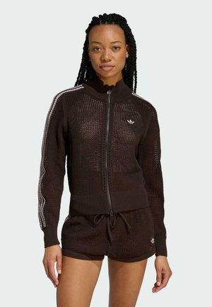 Frau mit geflochtenen Haaren, die eine schwarze Netzjacke mit Reißverschluss und passende Shorts mit weißen Adidas-Streifen und Logo vor hellem Hintergrund trägt.