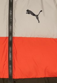 Kurtka puchowa z beżową górą, pomarańczowym środkowym fragmentem i ciemnozielonym dołem. Czarny zamek błyskawiczny z logo Puma na klatce piersiowej. Gładka faktura.