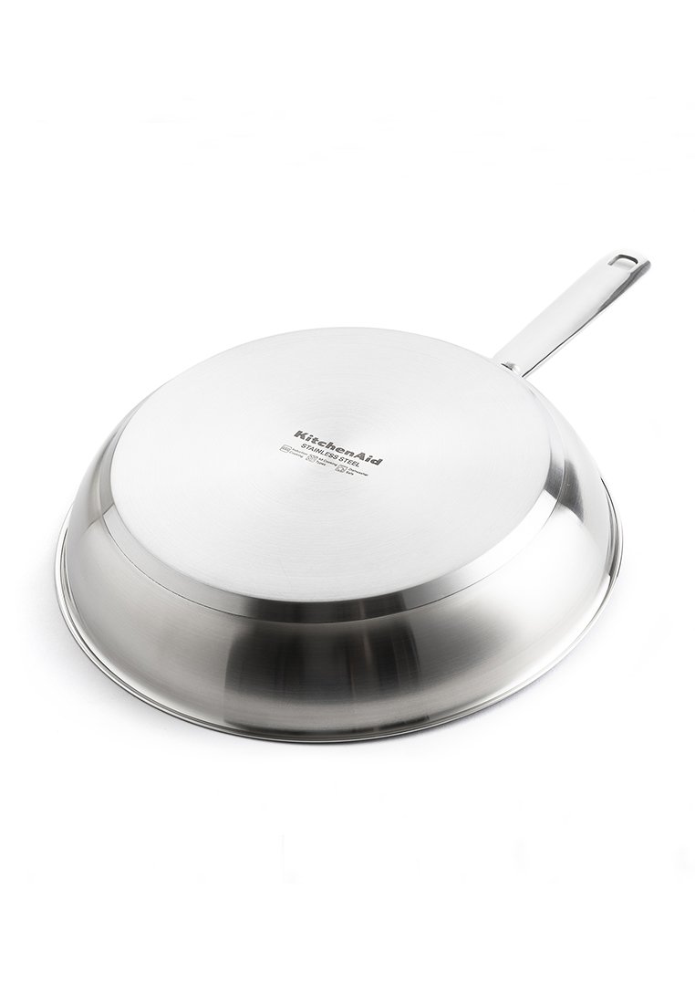 Patelnia ze stali nierdzewnej o gładkiej, błyszczącej powierzchni, zakrzywionym kształcie, rozszerzonym brzegu i długim, prostym uchwycie. Z logo KitchenAid.