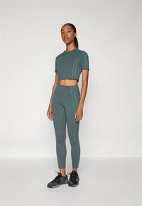 Grønt sett med crop top og leggings, med kontrasterende turkise sømmer og en tettsittende design. Brukt sammen med svarte treningssko. Glatt stofftekstur.