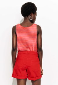 Shorts rouges avec des ourlets retroussés associés à un débardeur rayé corail et rose. Le tissu semble doux et a une coupe décontractée. Des boucles d'oreilles circulaires sont visibles.