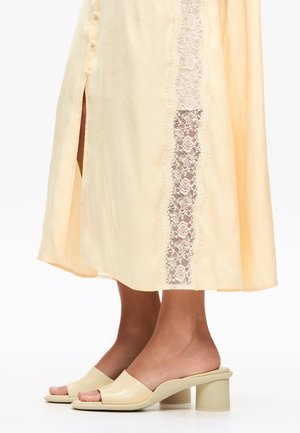 Jambes inférieures portant des sandales à talons crèmes et une robe jaune pâle avec une fente sur le côté et des détails en dentelle verticale.