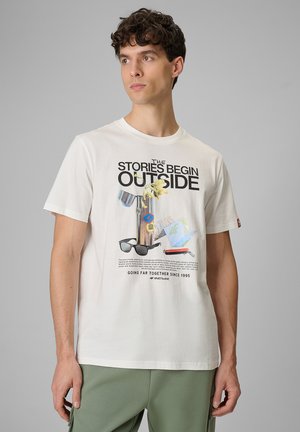 Młody mężczyzna noszący białą koszulkę z napisem „The stories begin outside” i nadrukiem sprzętu outdoorowego, stojący na jednolitym szarym tle.