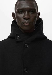 Abrigo de lana negro con cuello alto y grandes botones, presenta un diseño estructurado y textura suave. Adecuado para clima frío.