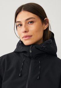 Zwarte hoodie-jack met een hoge kraag, voorzien van een ritssluiting aan de voorkant, verstelbare trekkoorden en een gestructureerde stofafwerking.