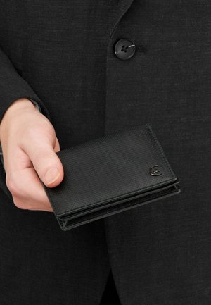 Ruka držící zavřenou černou peněženku s texturou na pozadí tmavé knoflíkové bundy.