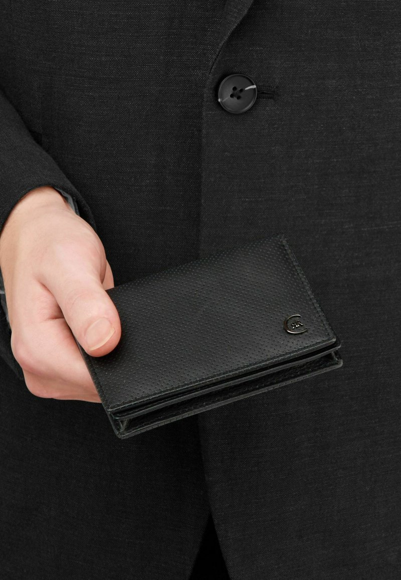 Mano sosteniendo una cartera negra texturizada cerrada sobre un fondo de chaqueta oscura con botones.