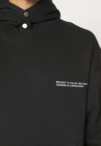 Sort hættejakke med en blød tekstur, der har en høj krave, knapper og hvid tekst: "BRAGT TIL DIG AF ORG DEPT® DESIGNET I KØBENHAVN."