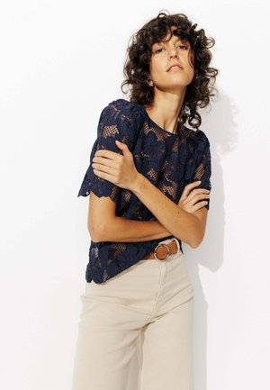 Top en dentelle bleu marine avec motif floral, manches courtes et texture transparente. Associé à un pantalon taille haute beige clair et une ceinture marron.