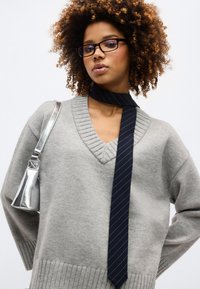 Νεαρή γυναίκα με σγουρά μαλλιά που φοράει μαύρα γυαλιά, γκρι πουλόβερ με V λαιμόκοψη, μαύρη ριγωτή γραβάτα και ασημί τσάντα ώμου σε λευκό φόντο.