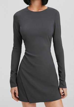 Woman wearing a fitted, long-sleeve, dark gray mini dress with a round neckline and subtle side drape on a plain background.