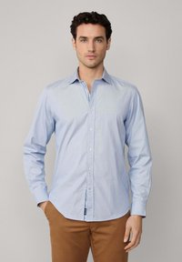 Camisa de manga larga a rayas azul claro con cuello abotonado, botones en el frente y una pequeña etiqueta con logo en el dobladillo; fabricada en suave algodón.