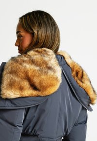 Giubbotto imbottito caratterizzato da un tessuto grigio scuro e un cappuccio in pelliccia sintetica in tonalità di marrone chiaro con striature più scure. Tra i dettagli è presente una cerniera.