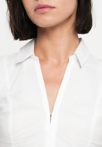 Λευκό πουκάμισο με γιακά και κλείσιμο με φερμουάρ, με λεία υφή και εφαρμοστή σχεδίαση. Ο γιακάς είναι καλοσιδερωμένος και καλοσχηματισμένος.