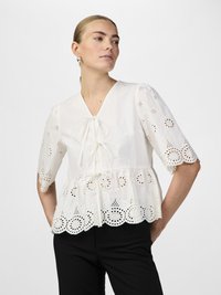Blusa bianca in cotone con scollatura a V, dettaglio legato, maniche corte e orlo in pizzo scallop con motivi floreali. Abbinata a pantaloni neri.