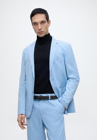 Άνδρας με ανοιχτό γαλάζιο κοστούμι και μαύρο ζιβάγκο με μαύρη ζώνη, στέκεται με το ένα χέρι στην τσέπη μπροστά σε απλό φόντο.