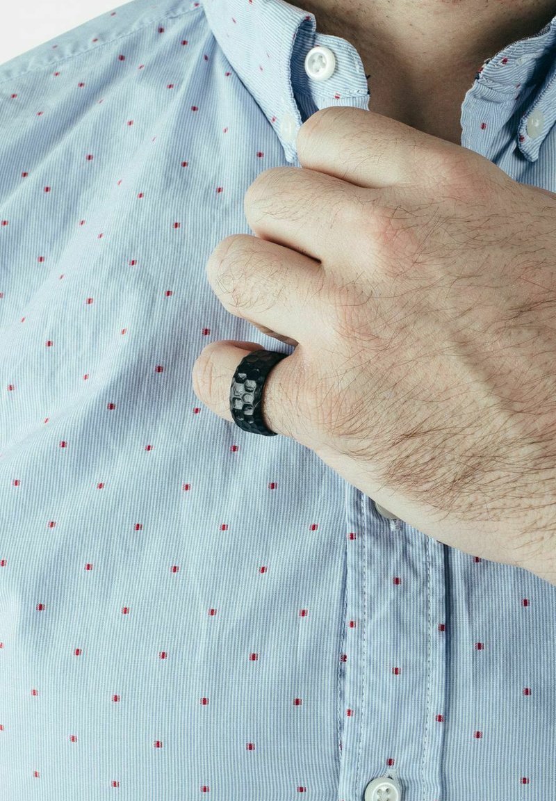Schwarzer texturierter Ring an einer Hand, getragen über einem blauen Hemd mit kleinen roten Punkten. Der Ring weist ein gehämmertes Muster und ein schlankes Design auf.