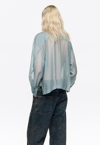 Jasnoniebieska, półprzezroczysta koszula z teksturowanym, kropkowanym wzorem i luźnym, swobodnym krojem. Posiada opadające ramiona oraz skrócony tył.