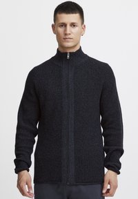 Suéter gris oscuro con textura, de cierre zip y cuello alto, con puños acanalados, que presenta un patrón de punto tejido y detalle de cremallera en la parte delantera.