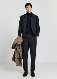 Completo slim fit di colore grigio scuro con finitura testurizzata, abbinato a un dolcevita nero. Un soprabito marrone appoggiato al braccio completa il look.