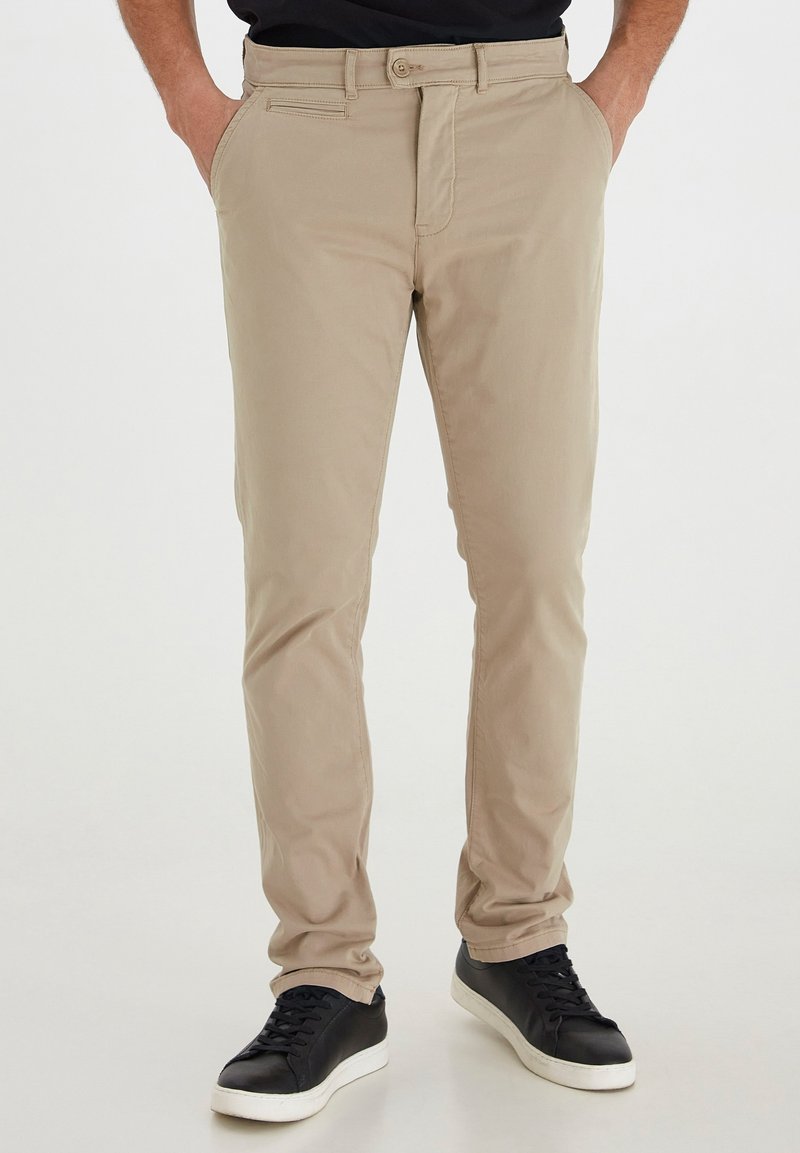 Homme portant un chino beige slim fit avec poches latérales et arrière, baskets noires avec semelles blanches, mains dans les poches.