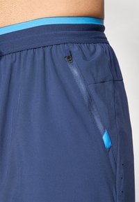 Närbild av marinblå träningsshorts med en sidoficka med dragkedja och detaljer i ljusblått runt midjan på en persons midja.