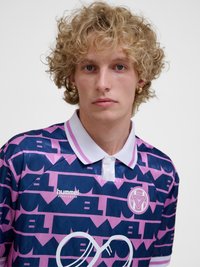 Młoda osoba z kręconymi blond włosami, ubrana w fioletową i granatową sportową koszulkę z białym kołnierzykiem i złotymi kolczykami kołami, zwrócona twarzą do przodu.