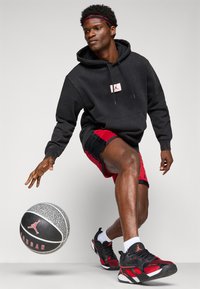 Zwarte hoodie met wit logo, zwarte shorts met rode accenten en zwart/rode sneakers, die een basketbal vasthouden, afgebeeld tegen een neutrale achtergrond.