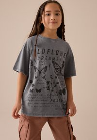 T-shirt gris oversize avec motifs de papillons noirs et texte. Manches courtes, encolure ronde et tissu doux. Design décontracté.