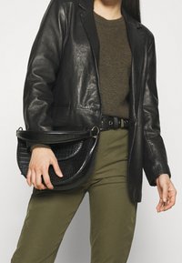 Borsa a tracolla in pelle nera con una texture a motivo coccodrillo, forma arrotondata e un manico singolo. Indossata con un outfit verde oliva.