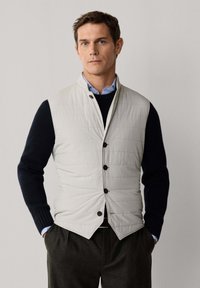 Homme portant une veste matelassée boutonnée gris clair par-dessus un pull noir et une chemise bleue à col, debout les mains dans les poches sur un fond uni.