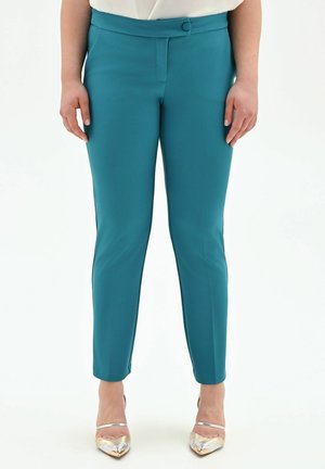 Donna con pantaloni su misura turchesi e décolleté metallizzati a punta, in piedi su sfondo bianco con le mani rilassate ai lati.