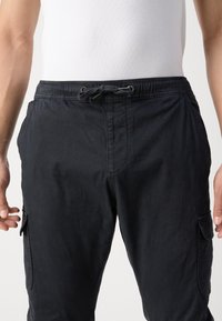 Zwarte cargobroek met een koordje in de taille, voorzien van zijzakken en een zachte, gestructureerde stof. Het ontwerp omvat accenten van cargozakken.