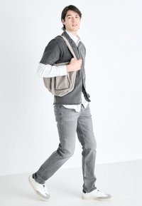 Jeans en denim gris, une chemise noire à manches courtes par-dessus une chemise longue à rayures blanches, et un sac messager beige. Marchant sur un fond blanc.