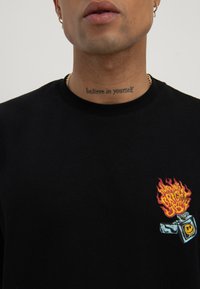 Close-up of a person wearing a black shirt with a lighter graphic and a "believe in yourself" neck tattoo.