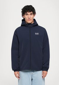 Fiatal férfi sötét, göndör hajjal, tengerészkék, zipzáras kapucnis dzsekit visel, amelyen az EA7 Emporio Armani logó látható, és világoskék farmernadrágot hord, fehér háttér előtt áll.