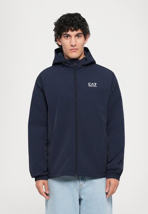 Ung mand med mørkt krøllet hår iført en navy zip-up hættejakke med EA7 Emporio Armani-logo og lyseblå jeans, stående mod en hvid baggrund.