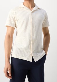 Korte mouwen, crèmewit shirt met een gestructureerd, wafelachtige patroon, knoopsluiting aan de voorkant en een klassieke kraag. Stof lijkt lichtgewicht en ademend.