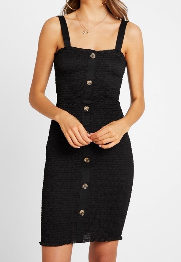 Kvinna i en svart texturerad ärmlös klänning med knappar framtill och en figursydd silhuett mot en vit bakgrund.