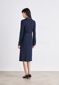 Navy ribbed dress with a collared neckline, long sleeves, and a belted waist, extending to mid-calf; paired with black heels.