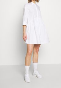 Abito di cotone bianco con una chiusura a bottoni sul davanti e maniche a tre quarti, caratterizzato da una vita arricciata e una gonna svasata, abbinato a sneakers bianche.