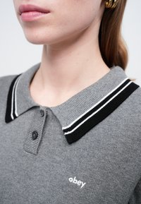 Pogled z bližine na osebo, ki nosi sivo pleteno polo majico s črno-belimi črtasto ovratnikom in logotipom "obey", pri čemer so vidni ustnice, vrat in zlat uhani iz obroča.
