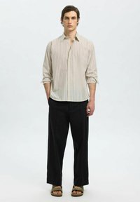 Camicia a bottoni di un leggero beige con righe verticali, abbinata a pantaloni larghi neri e sandali verdi. Design semplice e pulito.